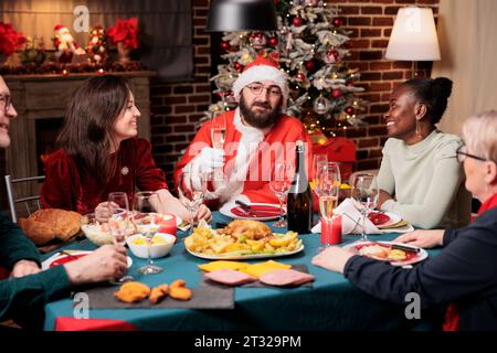 Il cosplay di Babbo natale diffonde positività intorno agli amici, si riunisce a tavola per celebrare l'evento della vigilia di natale. Un giovane adulto vestito con la tuta di babbo natale si sente allegro durante i festeggiamenti stagionali di dicembre. Foto Stock