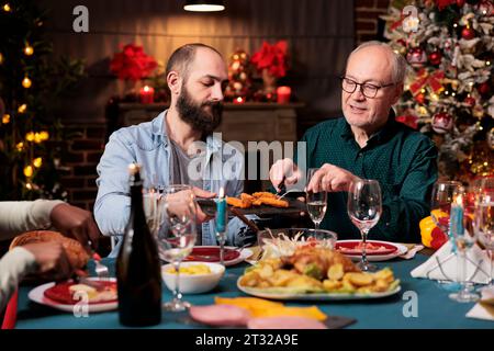 Persone diverse che si riuniscono intorno al tavolo per festeggiare le festività invernali, sentendosi felici durante la cena della vigilia di natale. Amici e familiari si godono il tempo a casa con piatti tradizionali fatti in casa. Foto Stock