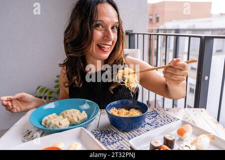 Attraente donna di mezza età che mangia cibo da asporto cinese seduto su un tavolo seduto in un balcone della città Foto Stock