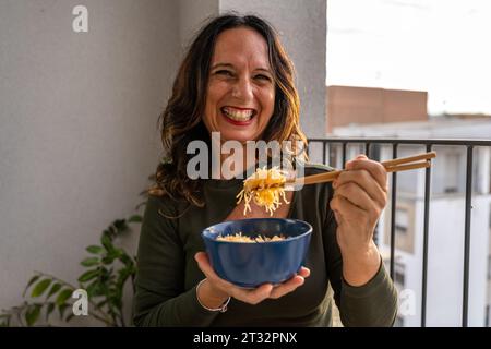 Attraente donna di mezza età che mangia cibo da asporto cinese seduto su un tavolo seduto in un balcone della città Foto Stock