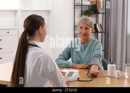 Giovane operatore sanitario che misura la pressione sanguigna della donna anziana al tavolo di legno all'interno Foto Stock