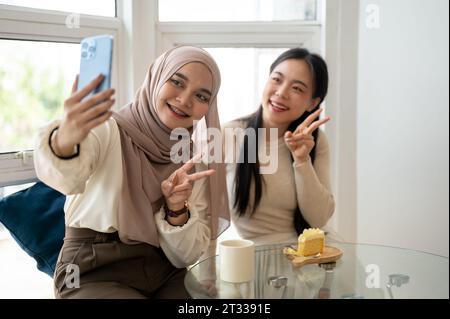 Una giovane donna asiatica-musulmana bella e felice sta facendo selfie con la sua amica mentre si gode in un bar. Foto Stock