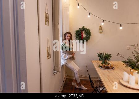Fiorista giovane donna in maglione a maglia bianca, seduta su davanzale, tenuta in mano il bouquet natalizio in carta artigianale in officina. Decorazioni invernali ecologiche Foto Stock