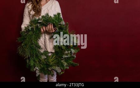 Banner. Giovane donna senza volto con maglione bianco a maglia tiene in mano la corona di Natale verde su sfondo scuro e isolato. Posiziona per testo. Nuovo Foto Stock