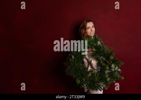 Giovane donna felice ed elegante con maglione bianco a maglia tiene in mano la corona di Natale verde su sfondo scuro e isolato. Posiziona per testo. Millen Foto Stock