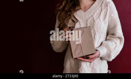 Banner. Giovane donna senza volto con maglione bianco a maglia regge il regalo della scatola in carta artigianale con corda bianca rossa legata alle mani su uno schienale isolato scuro Bordeaux Foto Stock