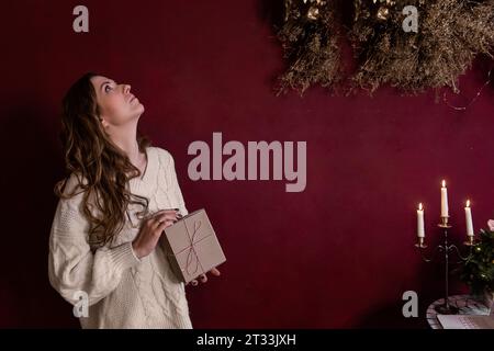 Elegante giovane donna con maglione bianco a maglia, racchiude una scatola regalo in carta artigianale con corda bianca rossa legata in mani su sfondo scuro isolato e Bordeaux. PLA Foto Stock