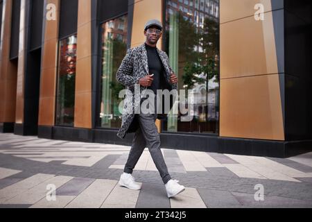 Bell'uomo che indossa abiti alla moda cappotto con stampa animale, dolcevita, berretto, pantaloni e occhiali. All'aperto. Model Model Walking City Street maschile ed elegante. Foto Stock