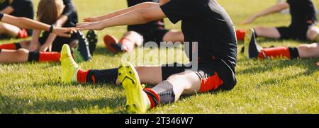 I bambini delle scuole si stendono sul campo sportivo in erba. Gruppo di giocatori di calcio in maglie nere di maglia da calcio che allungano le gambe dopo il treno Foto Stock