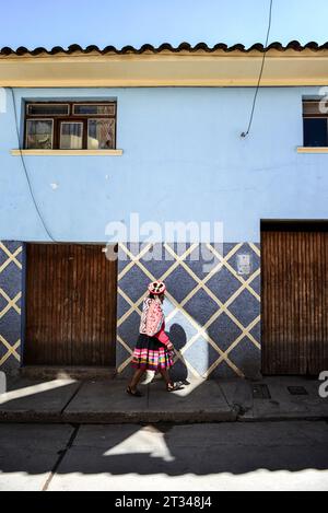 Una giovane donna del Quechuan cammina in un luogo soleggiato solitario in una strada ombreggiata a Urubamba. Foto Stock