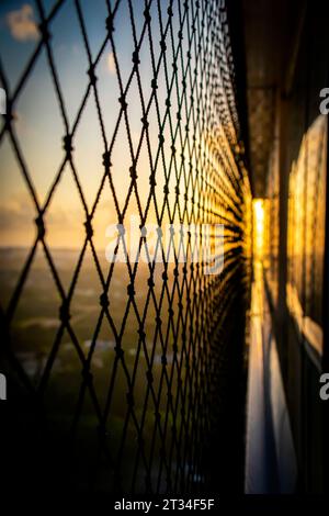 Il bagliore dell'alba che scorre attraverso una rete di sicurezza su un balcone, proiettando ombre intricate e una luce dorata soffusa sulla vista sottostante. Foto Stock