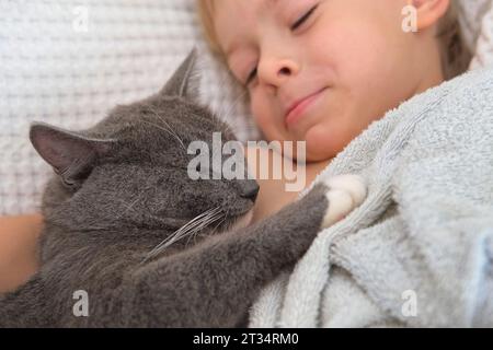 Ritratto del divertente piccolo ragazzo Cuddling Gray Kitten sdraiato in un letto. Concetto di animali domestici e bambini. Young Cat e Happy Smiling Kid dormono insieme a casa. Foto Stock