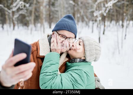 Donna anziana che bacia marito sulla guancia quando fanno selfie nel parco invernale Foto Stock