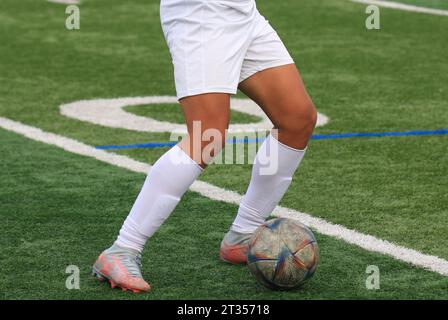 Primo piano delle gambe di un giovane giocatore di calcio in uniforme bianca con la palla e un campo in erba sintetica. Foto Stock