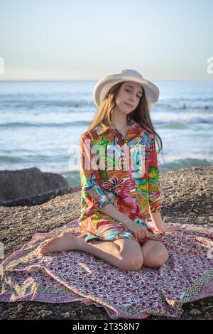 Nel tardo pomeriggio, una donna caucasica di 20 anni siede su un masso sulla spiaggia, adornata da un vivace abito da camicia a motivi geometrici e da un cappello elegante Foto Stock