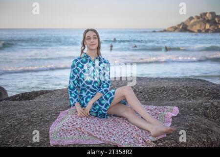 Nel tardo pomeriggio, una donna caucasica di 20 anni siede su un masso sulla spiaggia, adornata da un vivace abito camicia a motivi geometrici Foto Stock