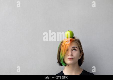 Ragazza adolescente con capelli tinti che bilanciano la mela sulla testa su sfondo grigio Foto Stock