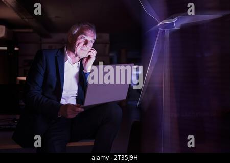 Uomo d'affari con un computer portatile che guarda alla futuristica turbina eolica in ufficio Foto Stock