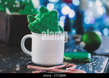 Biscotti smerigliati a forma di albero di Natale o biscotti con glassa verde all'interno di una tazza di caffè bianco con bastoncini di caramelle nelle vicinanze. Messa a fuoco selettiva con f. Sfocata Foto Stock