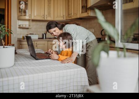 Madre che aiuta il figlio a guardare le lezioni online sul portatile al tavolo da pranzo Foto Stock