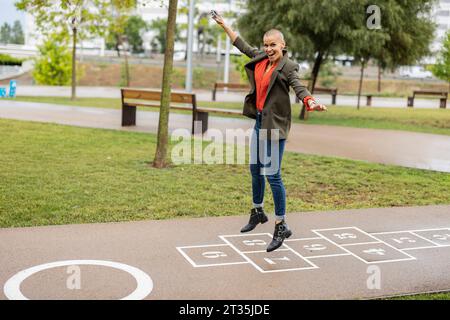 Donna spensierata con le braccia allungate che salta su un hopscotch al parco Foto Stock