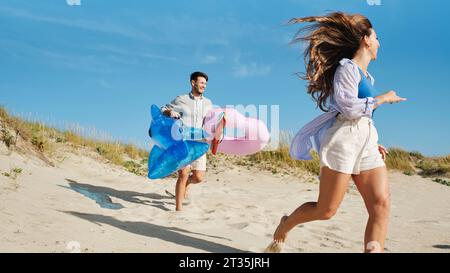 Donna felice che corre con un uomo che tiene l'anello gonfiabile e lo squalo giocattolo Foto Stock