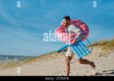 Uomo felice che corre con anello gonfiabile e sedia pieghevole in spiaggia nelle giornate di sole Foto Stock