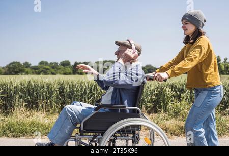 Ragazza sorridente con nonno seduto in sedia a rotelle e ascoltando musica nelle giornate di sole Foto Stock