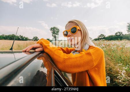 Donna che indossa gli occhiali da sole appoggiata all'auto Foto Stock