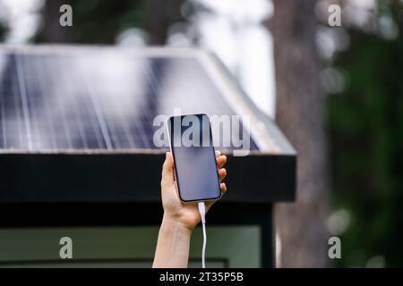 Mano di donna che tiene uno smartphone con pannello solare sullo sfondo Foto Stock