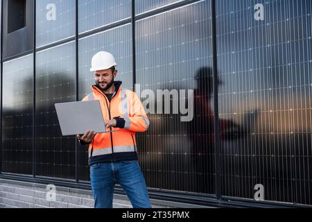 Ingegnere sorridente che utilizza un computer portatile in piedi vicino a pannelli solari Foto Stock