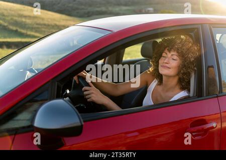 Donna sorridente con gli occhi chiusi che guida un'auto rossa Foto Stock
