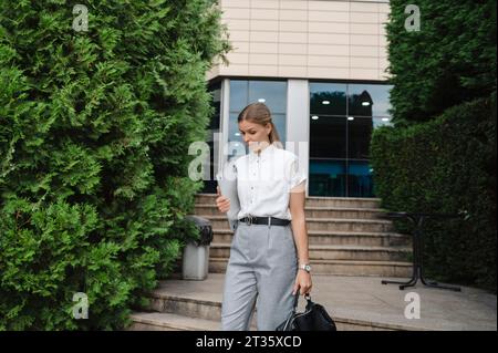 Donna d'affari che cammina con un computer portatile e una borsa davanti all'edificio Foto Stock