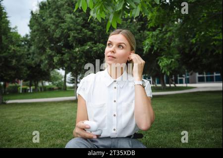 Donna d'affari con cuffie intrauricolari wireless seduto nel parco dell'ufficio Foto Stock