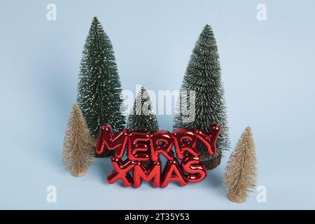 Un ornamento natalizio di plastica rosso lucido con un testo che dice allegri natali davanti a una foresta di alberi di natale dorati e verdi innevati. Minimalista, Foto Stock