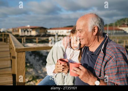 Felice coppia anziana che tiene tazze di caffè e si abbraccia nelle giornate di sole Foto Stock