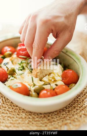 Mano di persona che mangia formaggio feta al forno con olive, pomodori ciliegini ed erbe Foto Stock