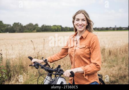 Donna sorridente che tiene una bicicletta in piedi vicino al campo Foto Stock