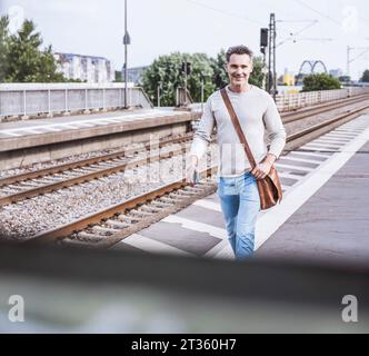 Uomo felice con la borsa a spalla che cammina alla stazione ferroviaria Foto Stock