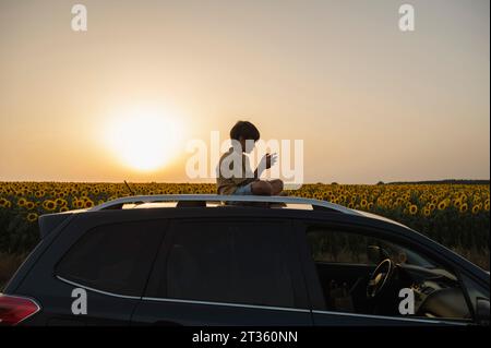 Ragazzo che usa uno smartphone seduto in cima all'auto al tramonto Foto Stock