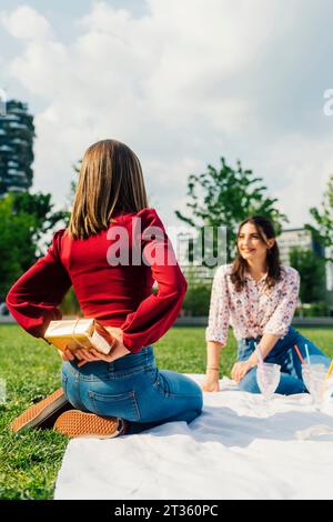 Donna che nasconde la confezione regalo dietro la schiena davanti a un amico sotto il cielo nuvoloso Foto Stock