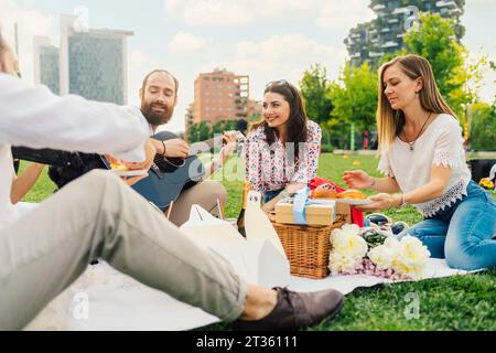 Uomo sorridente che suona la chitarra con gli amici e che si divertono a fare un picnic nel parco Foto Stock
