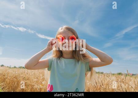 Ragazza sorridente che tiene i fiori sugli occhi nel campo di grano nelle giornate di sole Foto Stock