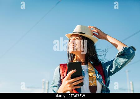 Donna felice che indossa un cappello e sta in piedi con lo smartphone sotto il cielo Foto Stock