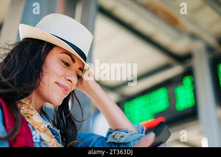 Donna sorridente che indossa un cappello che usa il telefono cellulare alla stazione del tram Foto Stock