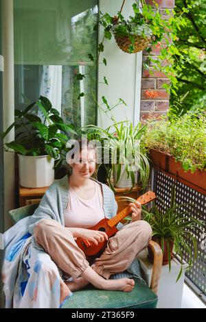 Adolescente sorridente con ukulele seduto su una sedia sul balcone Foto Stock