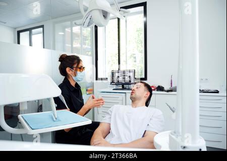 Dentista che indossa una maschera chirurgica che fornisce consigli al paziente nella sala esame Foto Stock