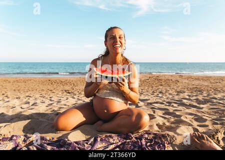 Donna felice incinta che mangia anguria sulla spiaggia Foto Stock