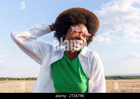 Donna afro felice che indossa cappello e occhiali da sole a forma di stella sul campo Foto Stock