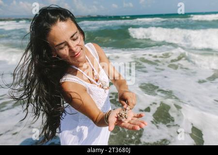 Donna con lunghi capelli che tiene conchiglie in mano vicino al mare in spiaggia Foto Stock